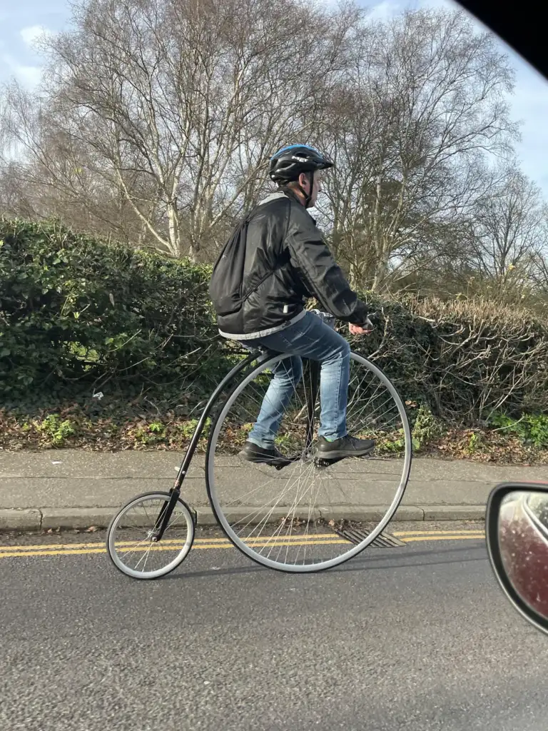 A person riding a vintage penny-farthing bicycle on a road lined with bushes and bare trees. The rider wears a helmet, a black jacket, and jeans. Part of a car's side mirror is visible in the foreground.