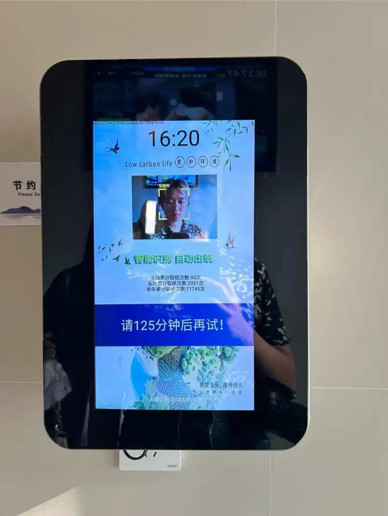 A digital kiosk display in a public place shows a message in Chinese and English about low carbon life. The screen includes an image of a person and a notification prompting the user to try again in 125 minutes.