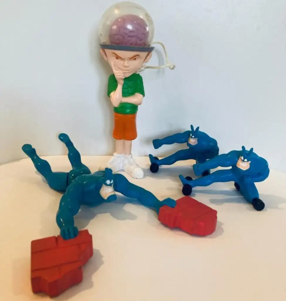 A figure with a transparent dome helmet, revealing a brain, stands with an evil smile and hand on chin. In front, three blue superhero figures are on their sides, two holding red blocks. The background is a plain light-colored surface.