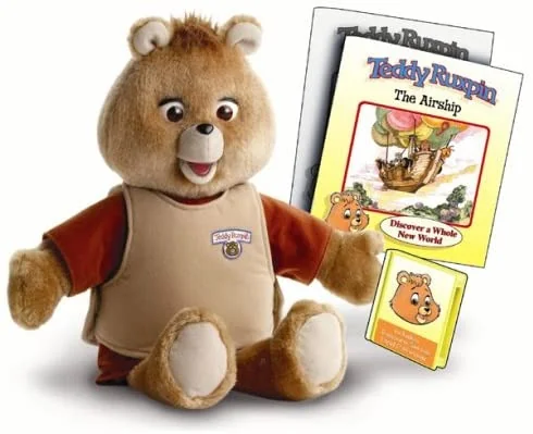 A Teddy Ruxpin bear sits beside storybook materials titled "The Airship." The bear is light brown with a beige vest and is positioned to face the viewer. The book cover features an illustration of an airship and a cartoon bear.