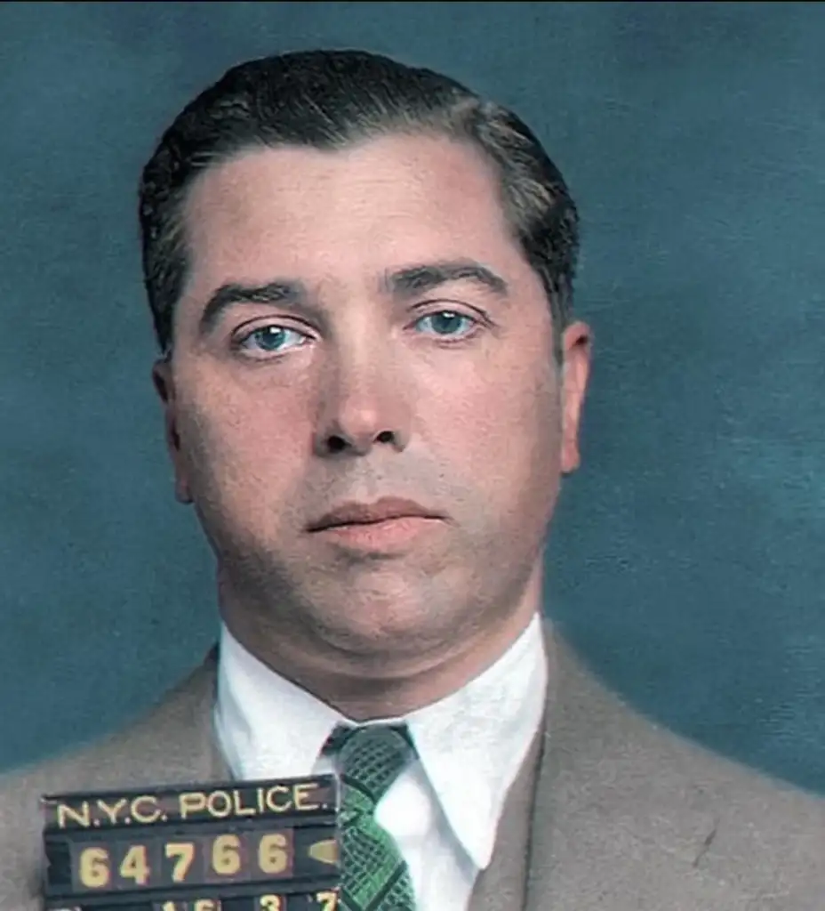 A vintage police mugshot of a man with dark hair and a neutral expression. He is wearing a suit with a patterned tie. The image is labeled with "N.Y.C. Police" and a number on a plaque.