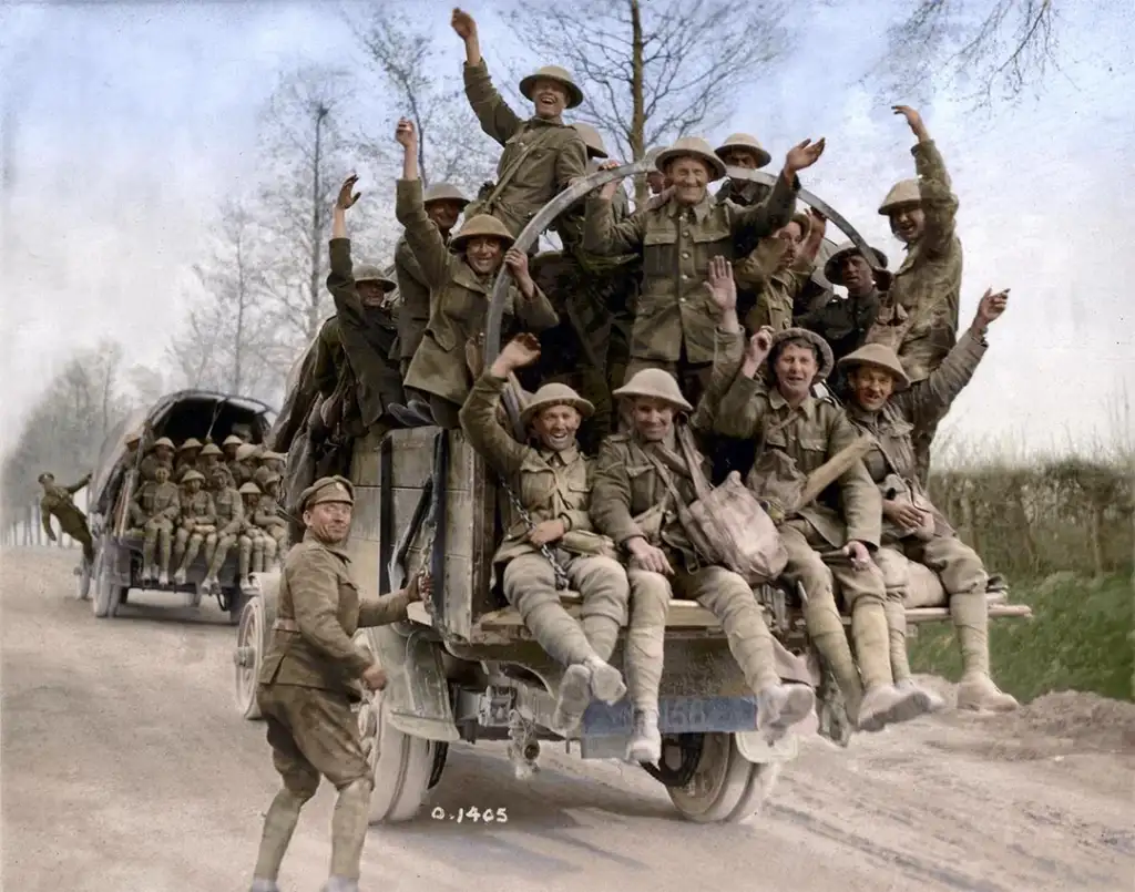 A group of soldiers in World War I uniforms are joyfully sitting and standing on a truck, raising their arms in celebration. Another truck full of soldiers is visible in the background on a rural road. Trees line the road.