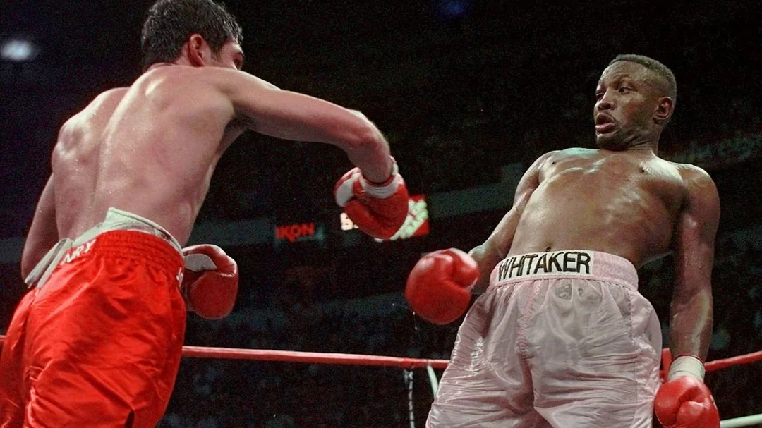 Two boxers in a ring: the boxer on the left in red trunks throws a punch, while the boxer on the right in white trunks and "WHITAKER" on the waistband leans back to evade it. The crowd is visible in the background.