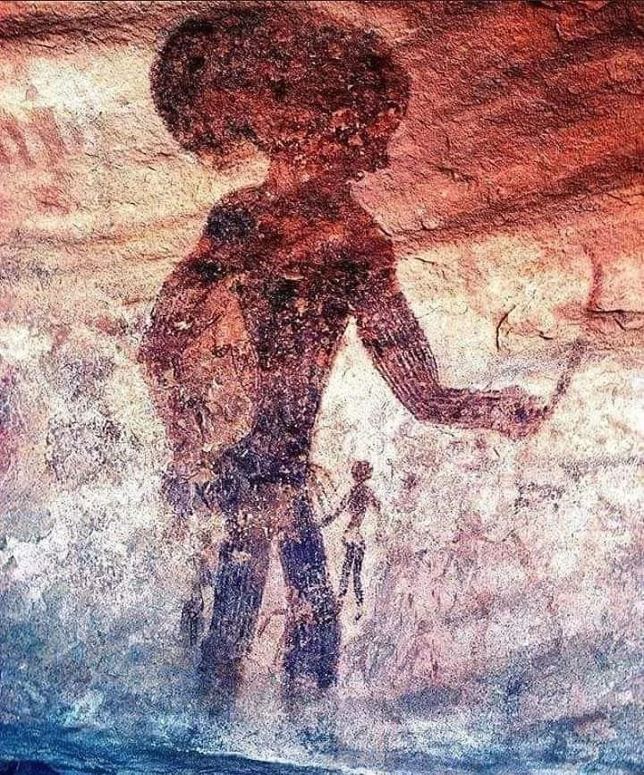 Ancient rock art depicting a large human figure with an elaborate hairstyle, holding an object. A smaller figure stands nearby. The artwork is on a reddish-brown stone surface, rich with texture and natural colors.