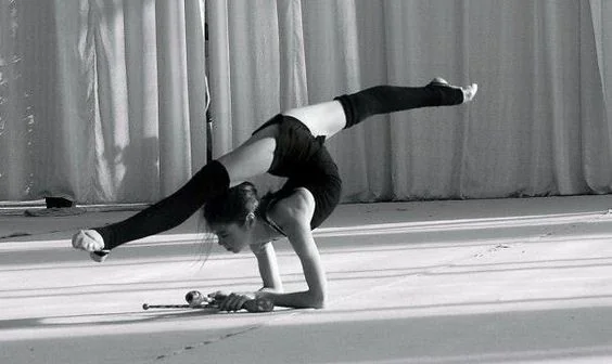 A person performing a balance pose on one hand, legs extended and elevated above the body. They appear to be on a stage with curtains in the background, conveying strength and flexibility.