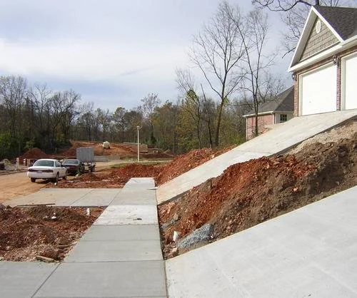 Steep driveways lead down from two garages, adjacent to newly laid pavements. A car is parked on a dirt road, surrounded by bare trees and construction materials. The sky is overcast.