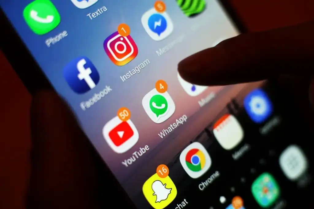 Close-up of a smartphone screen displaying various app icons including Facebook, Instagram, Messenger, YouTube, WhatsApp, Snapchat, and Chrome. A finger is about to tap the WhatsApp icon, which shows 4 unread messages.