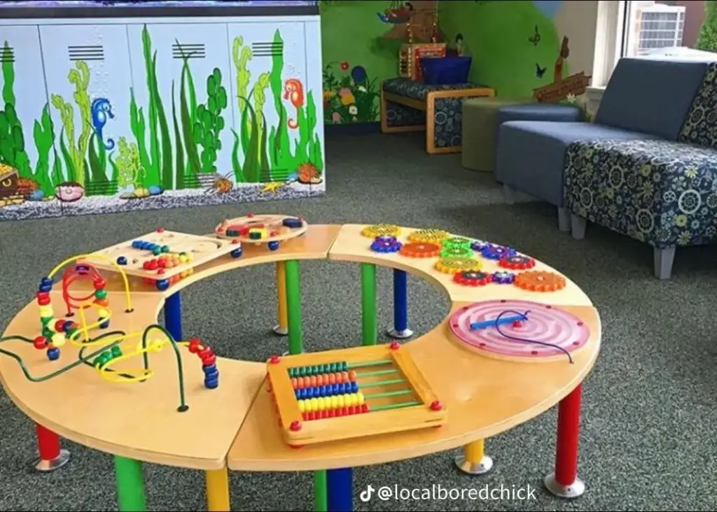 A colorful children's play area with a circular activity table featuring bead mazes, puzzles, and an abacus. The background has aquatic-themed wall decor, and there are blue patterned chairs nearby.