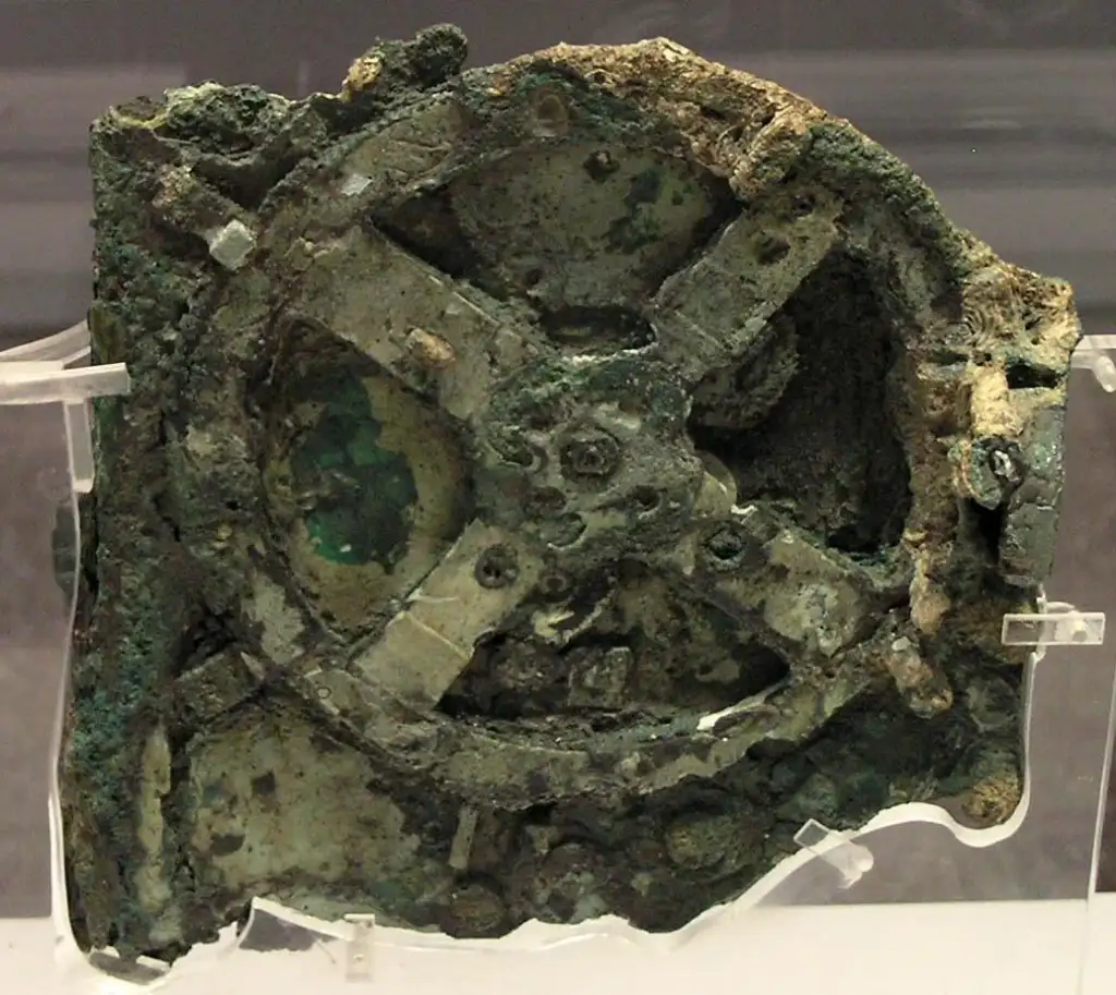 Rusted, ancient mechanical device with visible gears and a corroded, greenish-brown surface, displayed on a stand. The device appears weathered and partially broken, revealing intricate components.