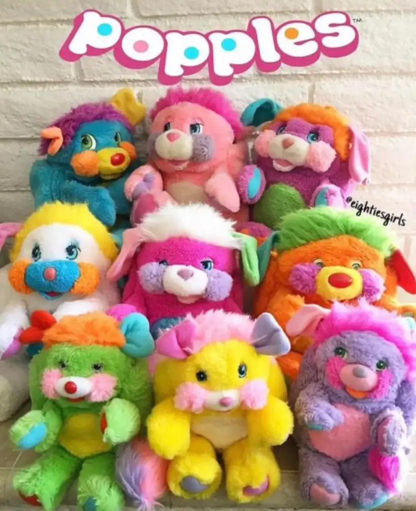 A group of colorful Popples plush toys arranged in three rows against a light background. Each toy is unique in color, featuring vibrant combinations like pink, orange, blue, and green, with playful faces and fluffy ears. The Popples logo is visible above.