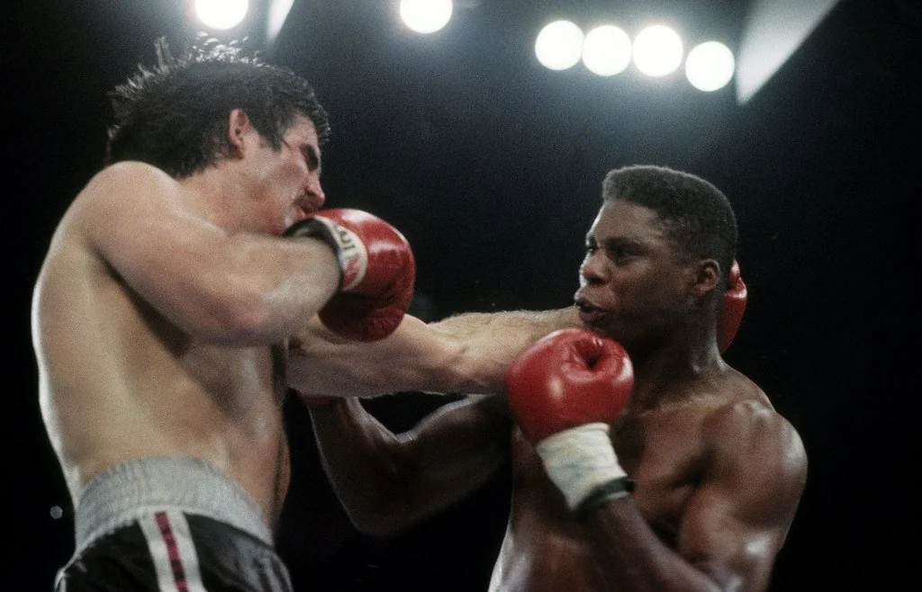 Two boxers in a ring, one delivering a powerful right punch to the other's face. Both wear red boxing gloves. Intensity and motion are captured mid-fight, with bright overhead lights illuminating the scene.