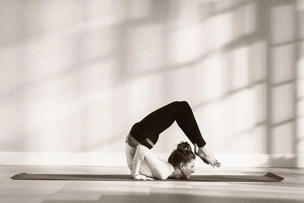 A person practicing an advanced yoga pose in a sunlit room, supporting their body on their forearms with legs bent over their back, feet touching near their head. They're on a yoga mat, and the background shows soft light patterns on the wall.