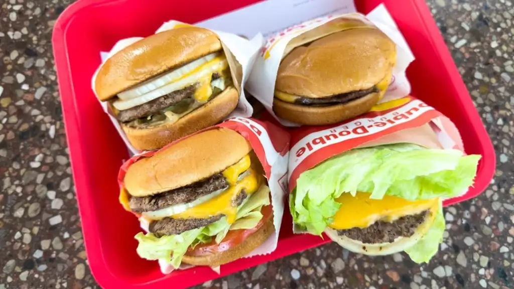 A red tray holds four burgers wrapped in branded paper. Each burger contains beef patties, melted cheese, lettuce, tomato, and onions, with a side of pickles. The tray is placed on a speckled surface.