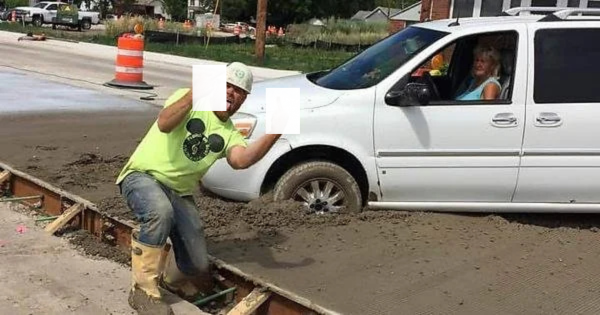 A construction worker in a green shirt and hat kneels on a newly paved road, gesturing with two covered fingers. A white van is stuck in the wet cement, with a woman driver inside, smiling. Construction cones are visible in the background.