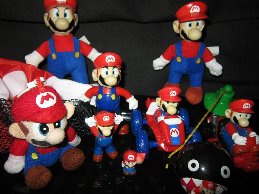 Various Mario-themed toys and figurines, including plush dolls and action figures, are displayed. A chain chomp toy and a Mario race car are also present. The characters wear red hats with an "M" logo and blue overalls.