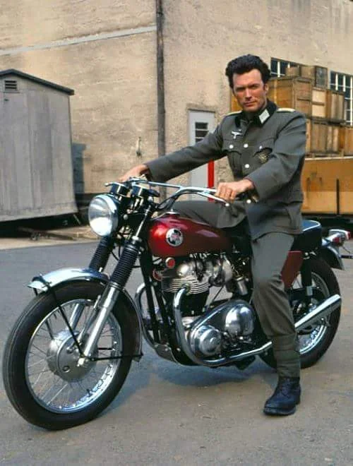 A man in a military-style uniform sits on a vintage motorcycle with a shiny, chrome finish and deep red accents. The background features a concrete wall, wooden crates, and a small shed. The scene conveys a sense of classic style and nostalgia.