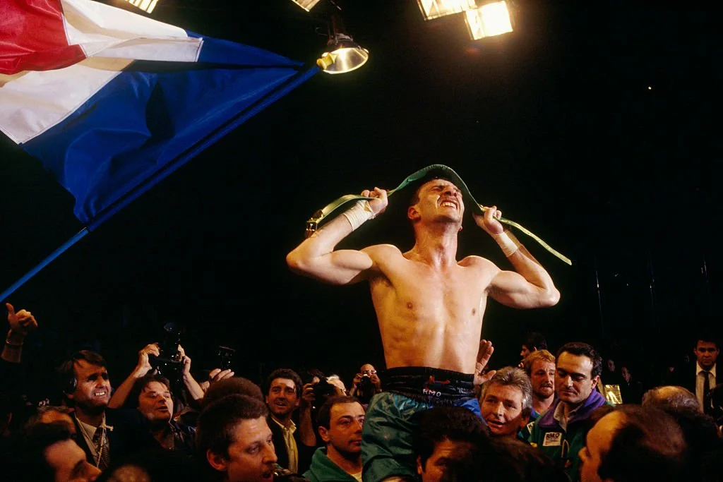 Boxer celebrating victory in a crowded ring, holding a green belt overhead. The French flag is held by someone nearby. He is surrounded by enthusiastic spectators and photographers under bright lights.