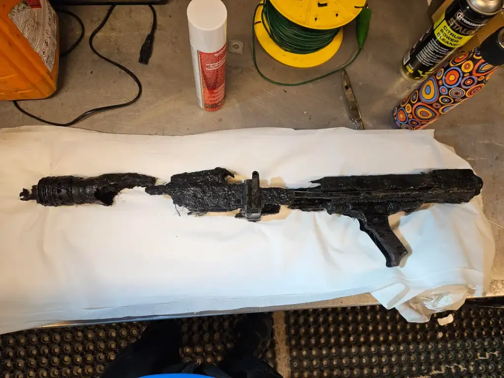 A severely charred, distorted airsoft rifle is placed on a white cloth on a table. Surrounding items include a can, a yellow reel, a colorful patterned object, and a corkscrew, conveying an industrial or workshop setting.