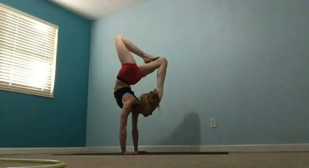 A person balances on hands in a yoga pose with legs arched over the back, touching feet to head. They are on a yoga mat in a room with light blue walls and a window with blinds.