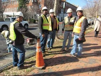 Five construction workers wearing safety vests and hard hats are gathered on a sidewalk near a street. One worker is placing an orange traffic cone, and others appear to be discussing something. Trees and buildings are visible in the background.