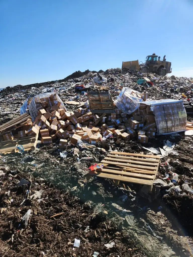 A large landfill site under a clear blue sky, with a bulldozer in the background moving piles of trash. Numerous cardboard boxes and wooden pallets are scattered across the heap of waste, creating a chaotic scene.