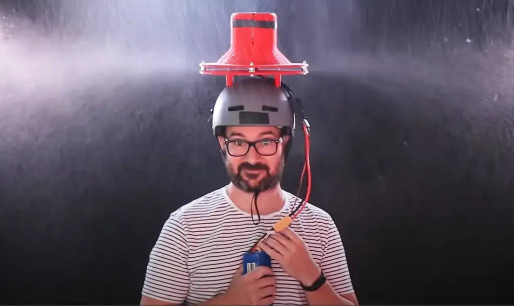 A person wearing a helmet with a red contraption on top, standing under a spray of water. They are holding a soda can and smiling at the camera. The background is dark, highlighting the water spray.