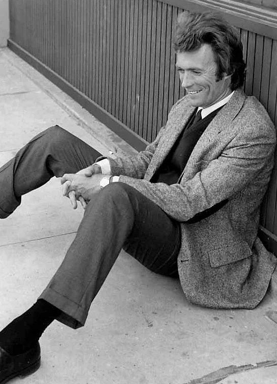 A man in a suit and tie is sitting on the ground against a wooden wall, smiling with his hands clasped around his knees.