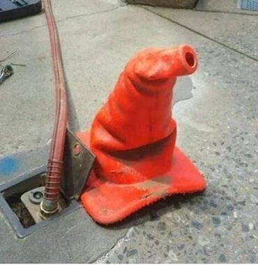 A red traffic cone shaped like a wizard's hat is placed on a concrete surface next to a partially exposed cable and a metal bracket. The cone appears bent and distorted.