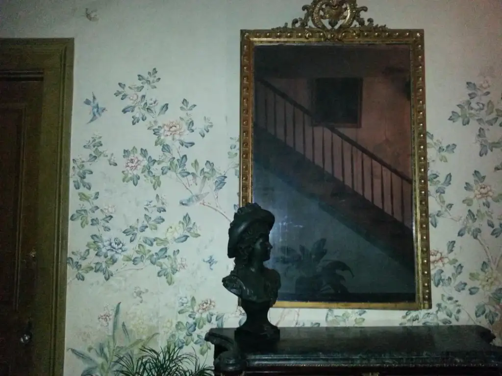 A decorative wall features floral wallpaper and a large, ornate mirror reflecting a staircase. Below the mirror, a bust sculpture is displayed on a dark table. The setting has an antique, elegant ambiance.