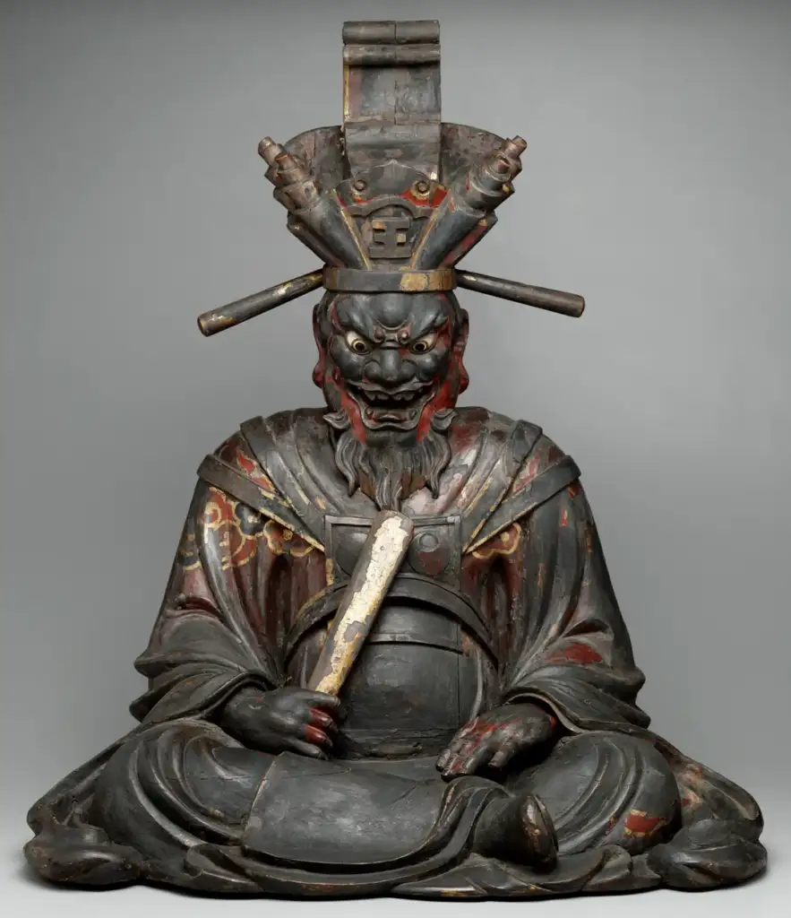 A wooden statue of a fierce-looking deity is seated, holding a scroll. It has an ornate headdress with rod-like extensions and wears a traditional robe with red accents. The expression is intense, with detailed facial features.