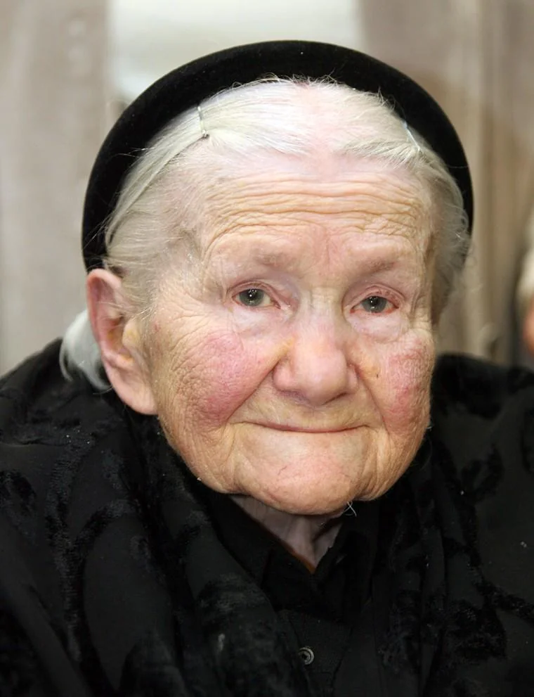 Elderly woman with white hair wearing a black headband and black clothing, looking into the camera with a gentle expression. The background is blurred.