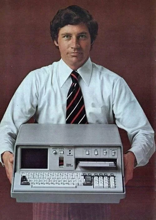 A person in a white shirt and striped tie holds an early IBM portable computer against a brown background. The computer features a small screen, keyboard, and mechanical buttons.