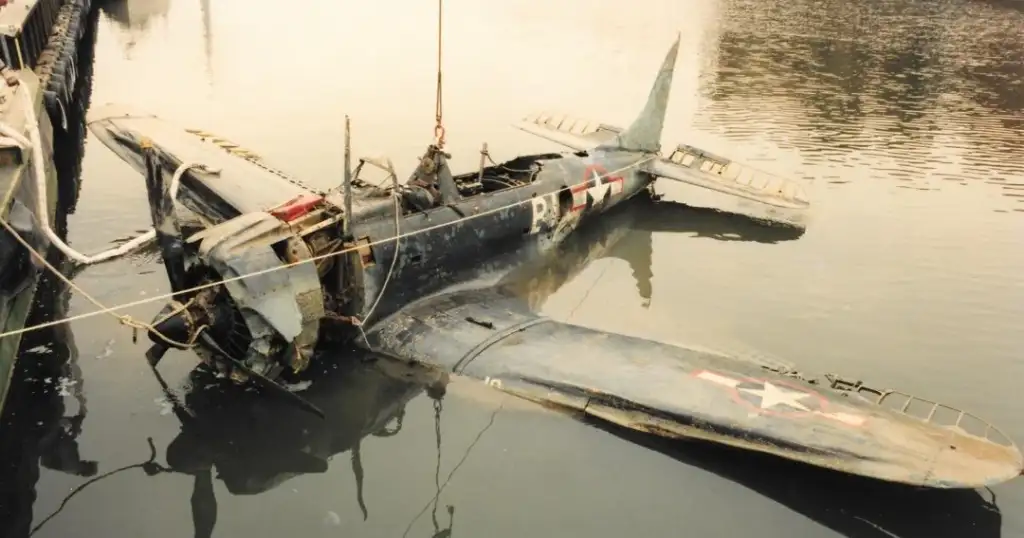 Partially submerged, damaged World War II-era fighter plane being lifted from water. The aircraft has visible wear, a missing propeller, and U.S. military insignia. Nearby ropes and a small crane are used for the recovery.