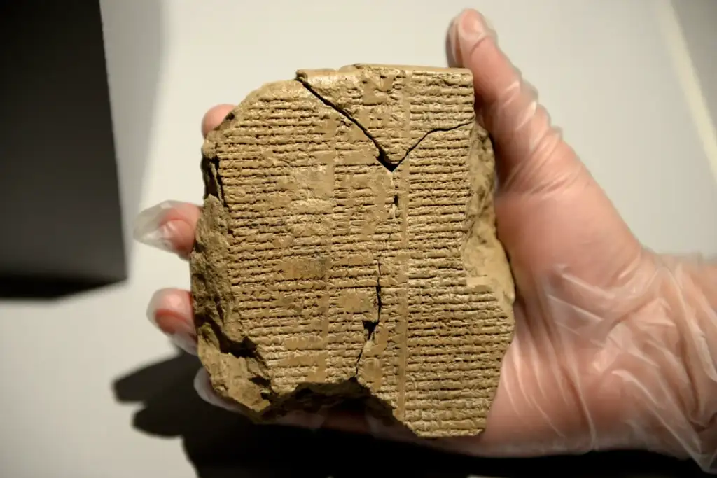 A hand in a glove holds a cracked, ancient clay tablet with intricate cuneiform script on its surface, set against a neutral background.