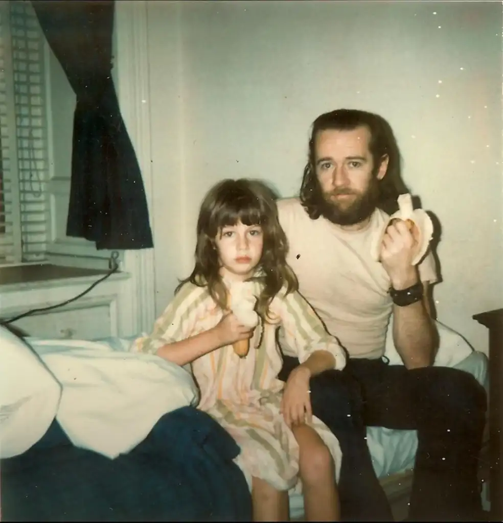 A man with long hair and a beard sits on a bed next to a young girl. Both are eating bananas. The room has a window with a dark curtain, and there is a blue and white blanket on the bed. The atmosphere appears casual and relaxed.