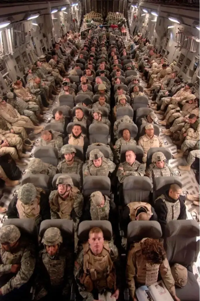 A large group of military personnel in uniform sit closely together in rows of seats inside an aircraft. The seats and interior are designed for transport, with equipment stored above. The atmosphere is orderly and focused.