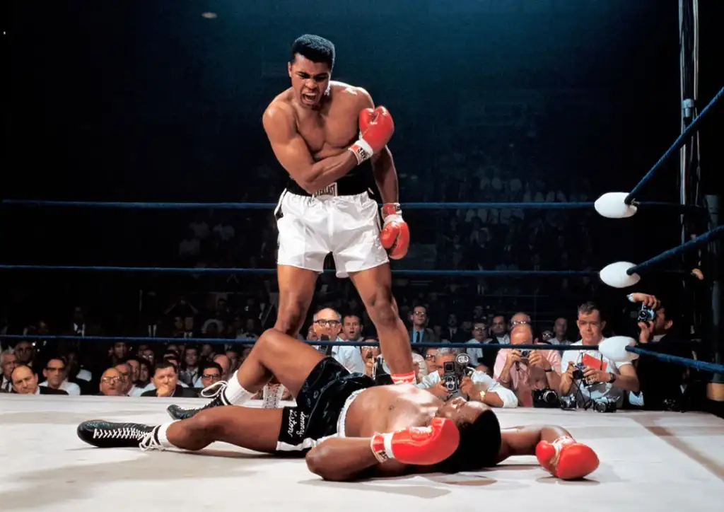 A boxer wearing white trunks and red gloves stands over an opponent lying on the canvas in a boxing ring. The standing boxer is shouting down at the fallen fighter. The scene is surrounded by onlookers and officials.