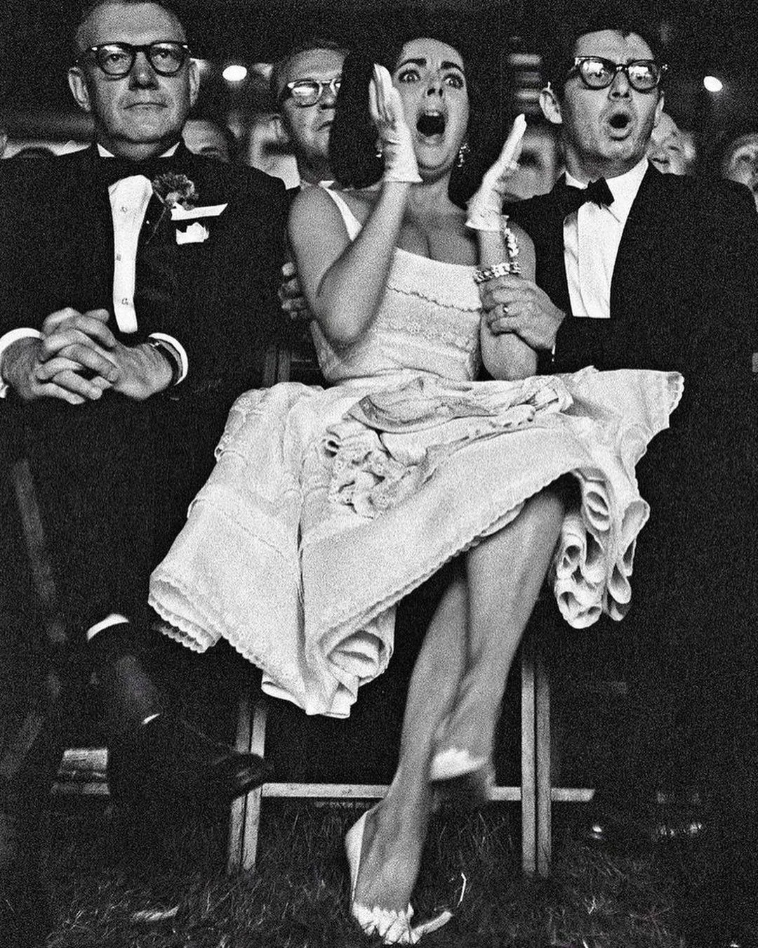 A black-and-white photo of three people sitting in a row. The woman in the center is clapping with an enthusiastic expression, wearing a 1950s-style dress. The two men beside her are wearing suits and glasses, with one crossing his arms.