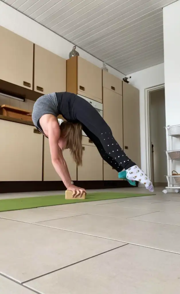 A person performs a backbend yoga pose in a kitchen, balancing with their hands on a yoga block. They wear a gray tank top, black leggings, and colorful socks. The kitchen has wooden cabinets, and a cart is visible in the background.