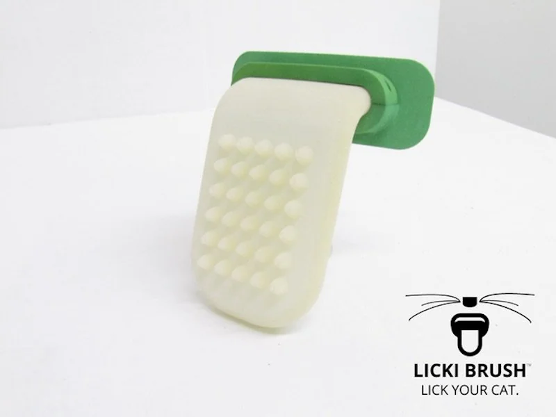 A green and white Licki Brush, a cat grooming tool, is displayed on a white surface. The brush has a handle and a textured area for grooming. The text "LICKI BRUSH: LICK YOUR CAT." and a cat face logo are visible.