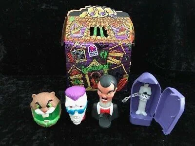 A set of Goosebumps-themed toys from Taco Bell is displayed. It includes a gopher, a blue skull with purple hair, a ventriloquist dummy, and a skeleton in a purple coffin. Behind them is a colorful box with Goosebumps branding.