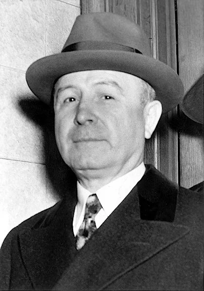 An older man wearing a suit and tie, with a fedora hat, stands against a wall, looking slightly to the side. The setting appears to be indoors and formal.