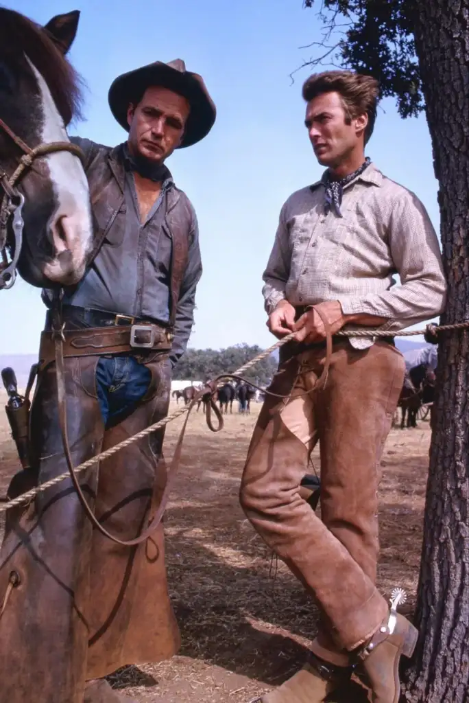 Two cowboys stand outdoors next to a horse. Both wear cowboy hats, shirts, and chaps. One leans against a tree with his legs crossed. A group of horses can be seen grazing in the background.