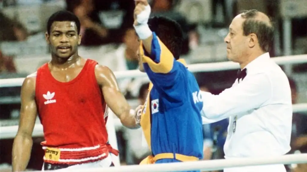 Two boxers and a referee in a boxing ring. The boxer in red looks on as the referee raises the arm of the boxer in blue and yellow, indicating victory. The setting appears to be a competitive match.