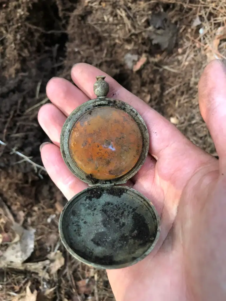 A hand holds an old, rusty pocket watch with its cover open. The watch is tarnished, showing signs of age and oxidation. The background is a forest floor with dried leaves and soil.