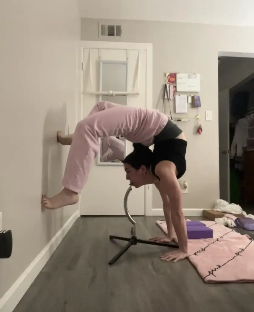 A person performs a yoga pose using a chair for support, balancing with legs arched over their back and feet pressed against the wall. They are in a room with wood flooring, pink yoga mat, and white walls.