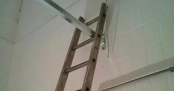 A long wooden ladder is precariously balanced against a wall, extending towards an unsecured tile ceiling. The setup appears unstable, suggesting a risky situation for anyone attempting to use it.