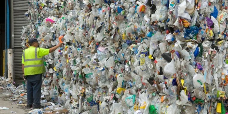 A worker wearing a high-visibility vest stands next to a massive pile of compacted plastic waste at a recycling facility. The pile consists of various plastic items, displaying a mix of colors and shapes.