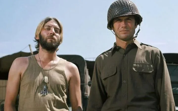 Two men stand side by side; one in a sleeveless shirt with a necklace, and the other in military attire with a helmet. They are outdoors in front of military vehicles, under a clear blue sky.