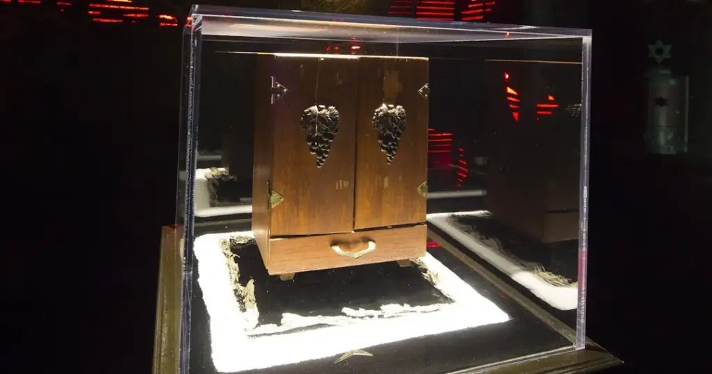 A wooden box with ornate carvings on its front, encased in a clear display case. The case has a white base, and the background is dimly lit with red accents.